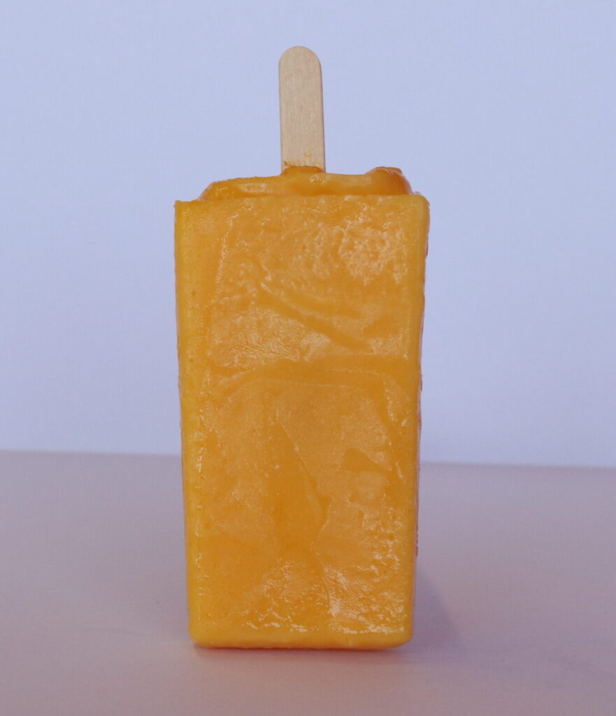 Bright yellow mango paleta standing upright with a wooden stick on a light surface