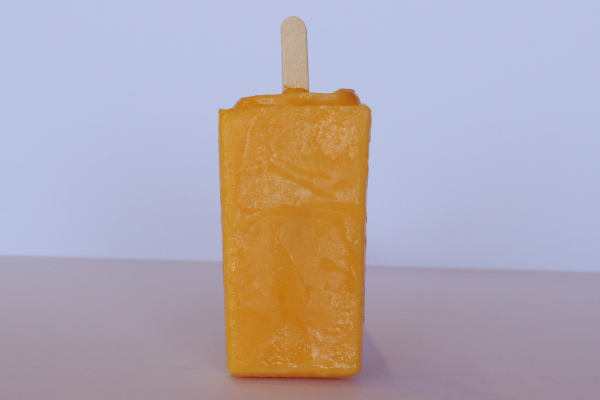 Bright yellow mango paleta standing upright with a wooden stick on a light surface
